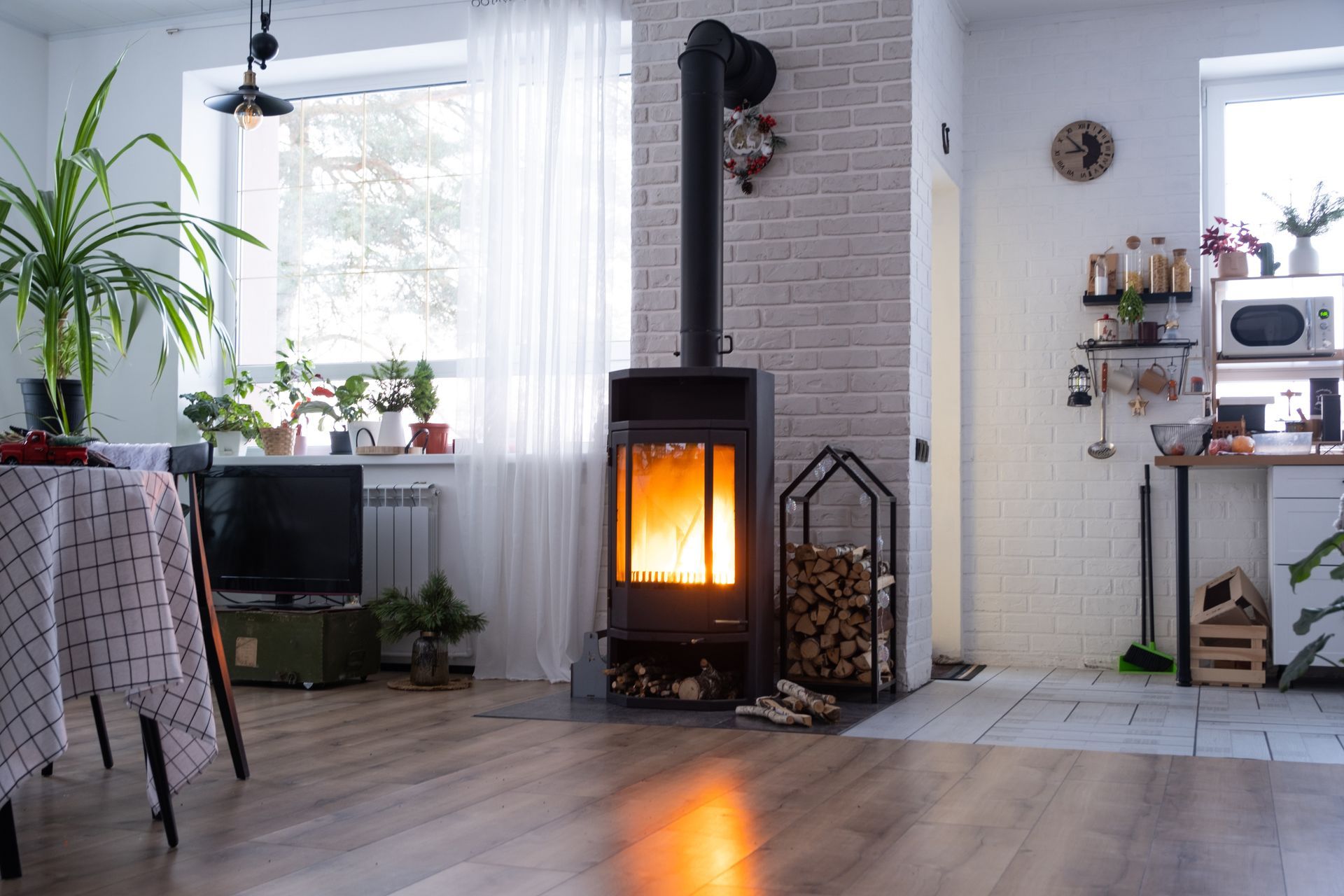 Cozy living space with lit fireplace against white brick, firewood nearby, window in background, light wooden floors.