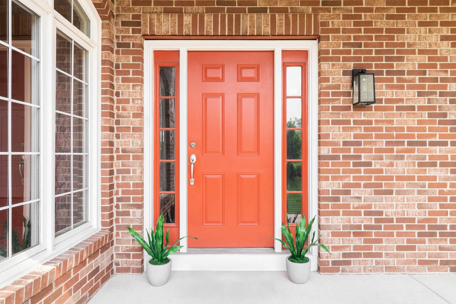 Red front door flanked by sidelights on a brick facade with a window, and potted plants.
