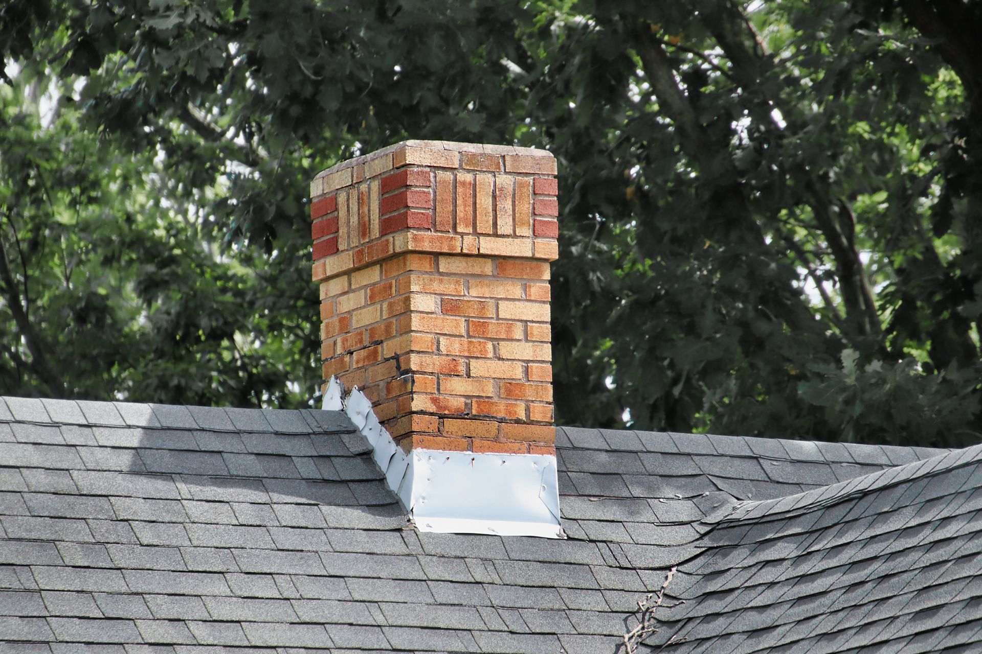 Brick chimney on a gray shingled roof, trees in the background.