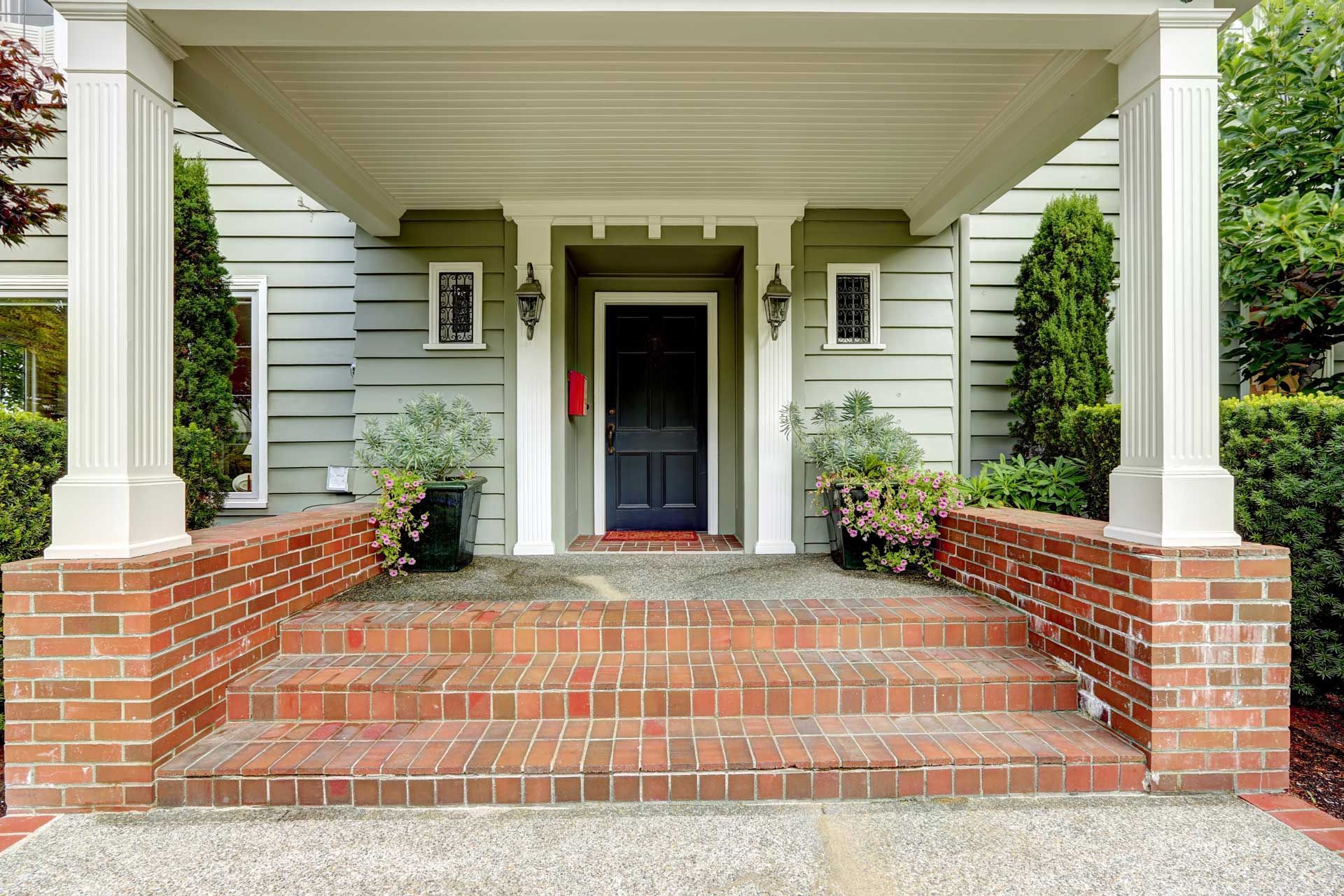 Brick steps leading to a house with a front porch. White columns frame the entrance, green siding, and a black door.