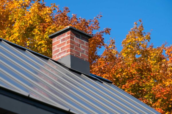Brick chimney on a metal roof with autumn foliage and a blue sky in the background.