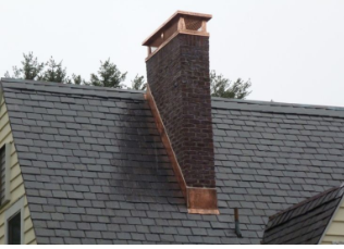 Brick chimney with copper flashing and cap on a gray shingle roof.