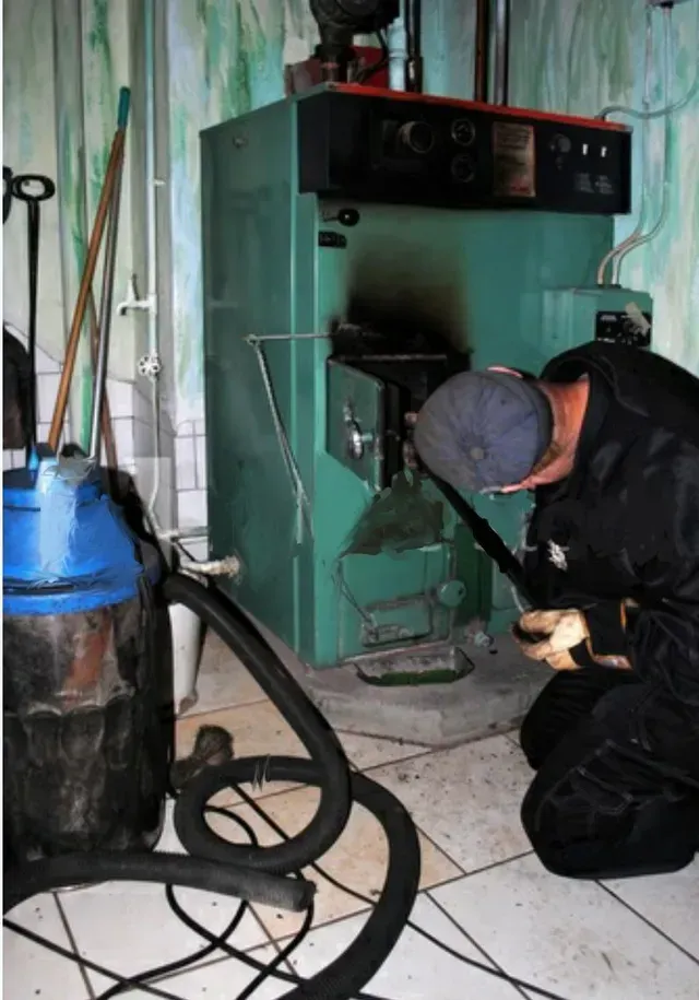 Person cleaning a large green boiler in a utility room, using a long tool and vacuum cleaner.