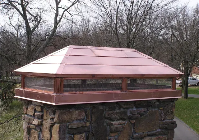 Copper chimney cap on a stone chimney. Trees and sky in background.
