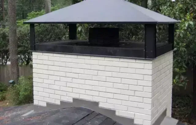 Brick chimney with black cap and metal hood on a rooftop surrounded by green trees.