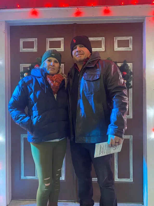 Couple posing in front of a decorated door with red and white lights. Woman in a puffer jacket and green pants. Man in a leather jacket.