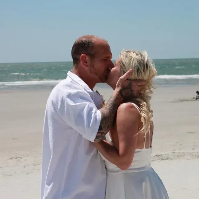 Couple kissing on a beach. The woman wears a white dress and the man has a tattoo on his arm.