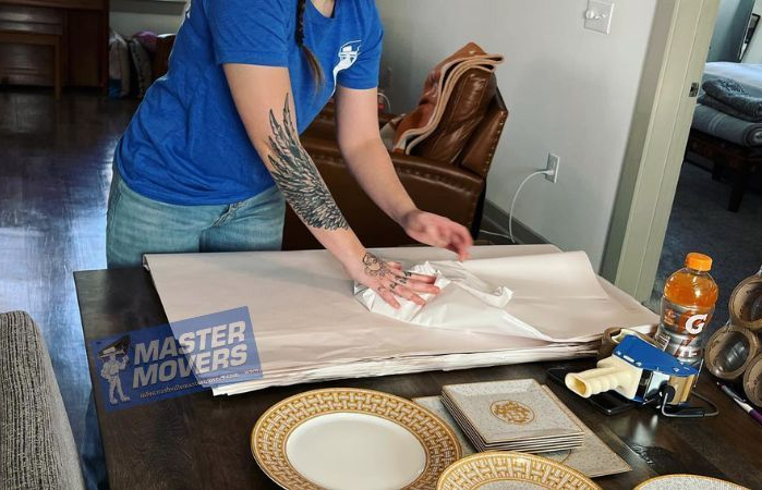A person in a blue shirt wrapping dishes on a table for moving, next to packing supplies.