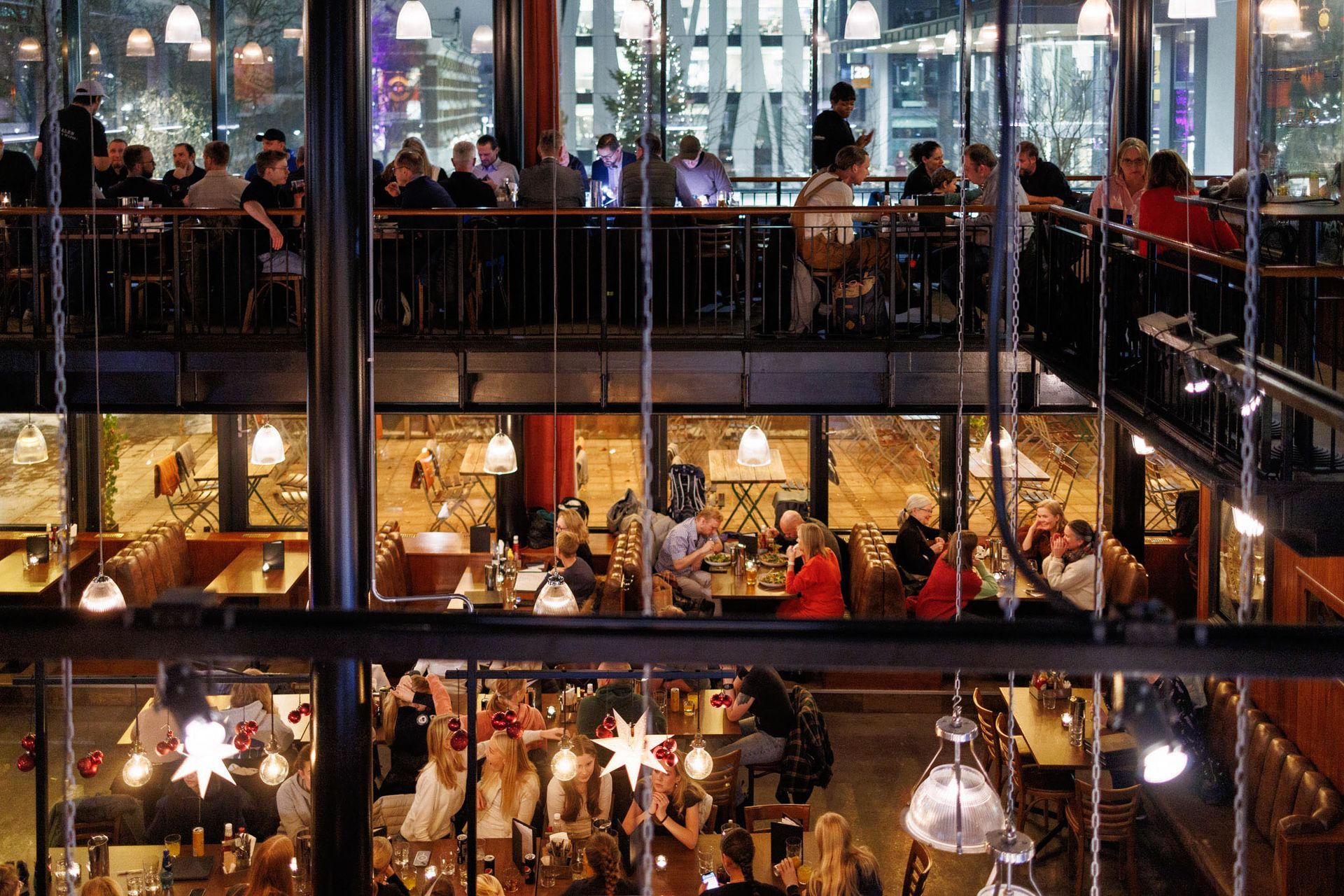Restaurant i flere etasjer, folk spiser. Store vinduer, metallrammeverk, varm belysning, sitteplasser i ulike nivåer.