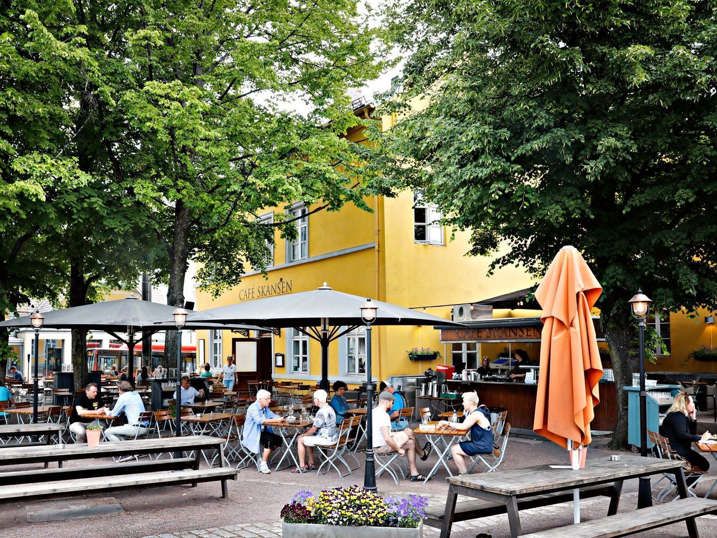 En gruppe mennesker sitter ved bordene under parasoller i en park.