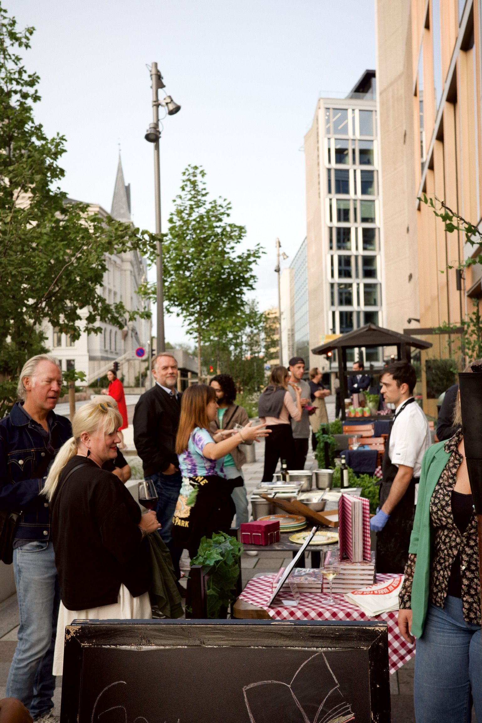 Folk på et utendørs matmarked, kvinne peker, selgere, bygninger i bakgrunnen.