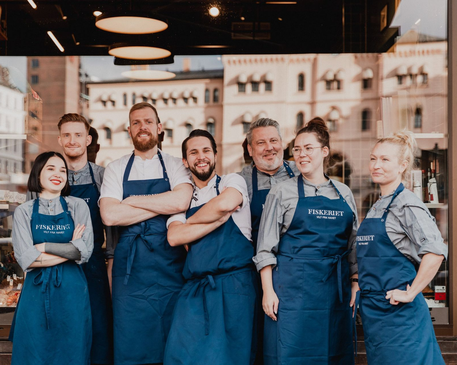 En gruppe restaurantansatte i blå forklær står foran en bygning.