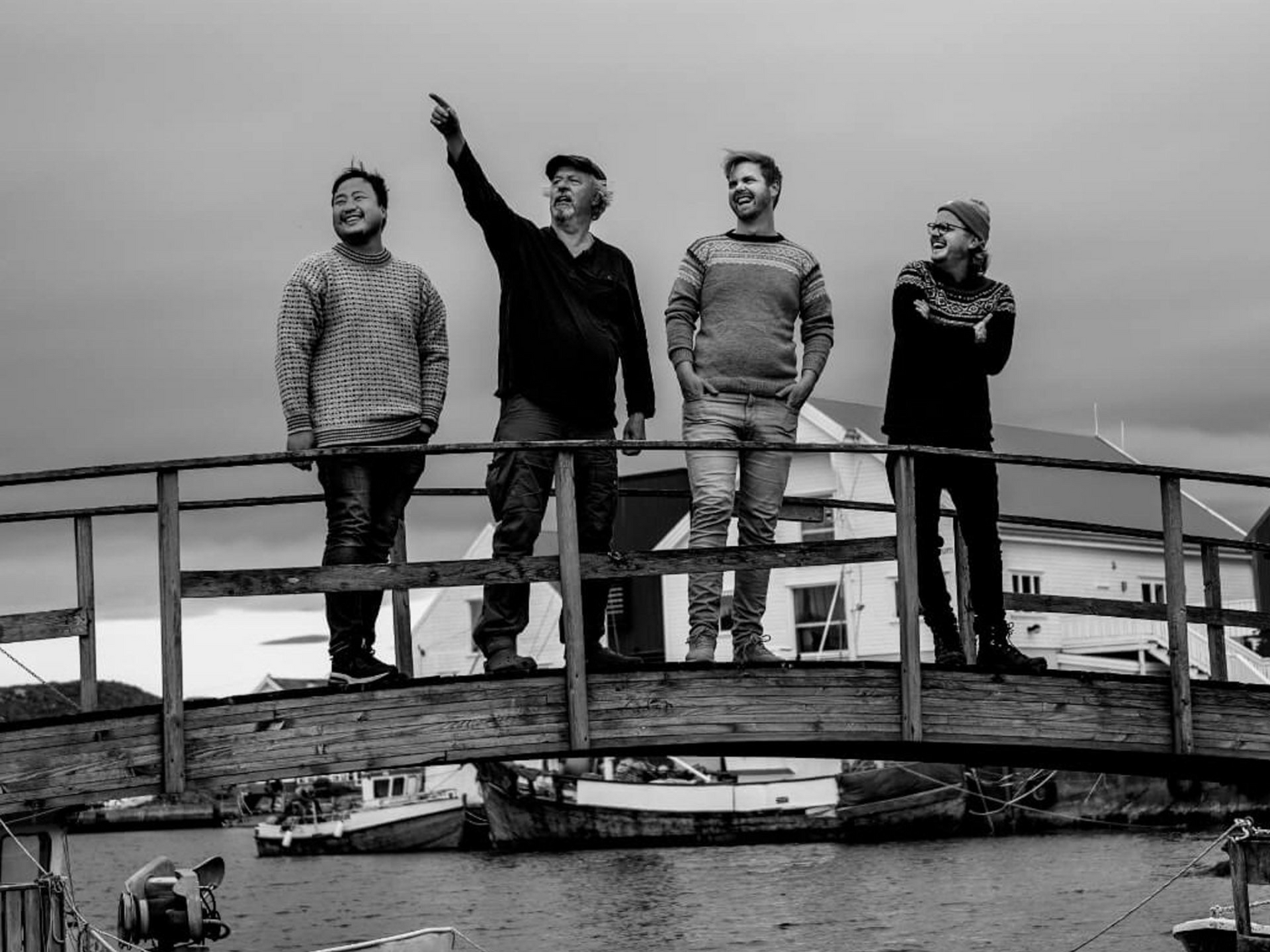 Fire personer på en trebro, én peker oppover. Overskyet himmel, vann og båter i bakgrunnen.