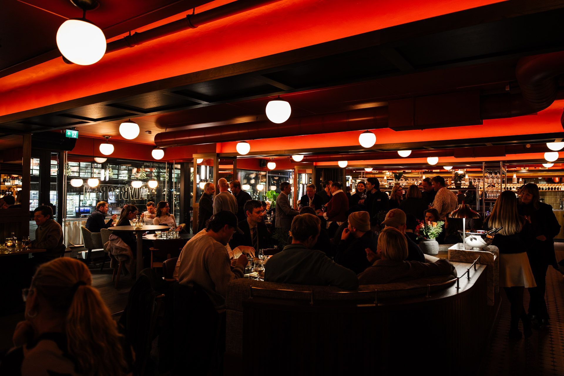 Restaurantinteriør med gjester sittende ved bord; rød belysning lyser opp taket.