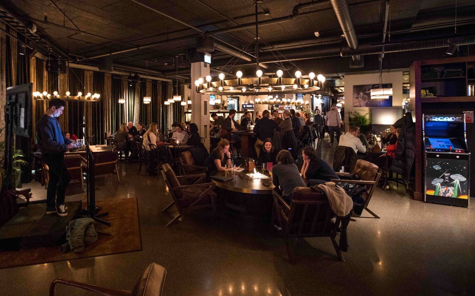 Folk i en svakt opplyst bar, noen sitter ved bord, andre står i nærheten av baren og arkadespill.