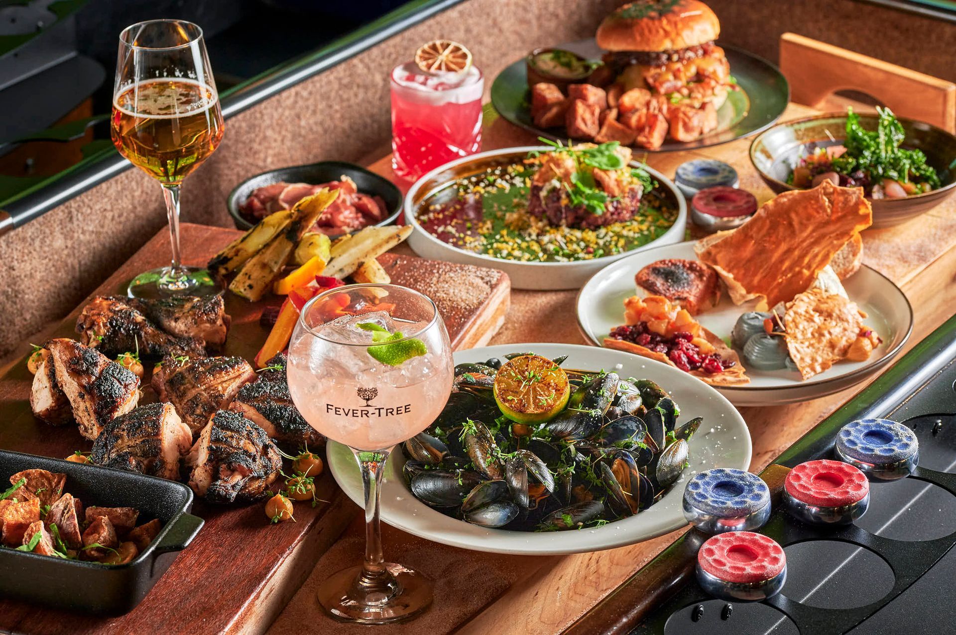 Et utvalg av mat og drikke på et bord, inkludert burger, blåskjell, cocktails og en øl.