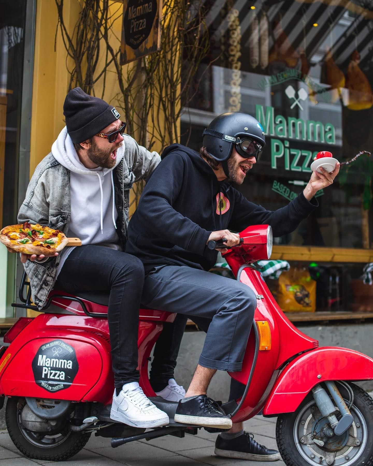 To menn på rød scooter, den ene holder pizza, den andre holder en liten dessert, foran butikken «Mammina Pizza».
