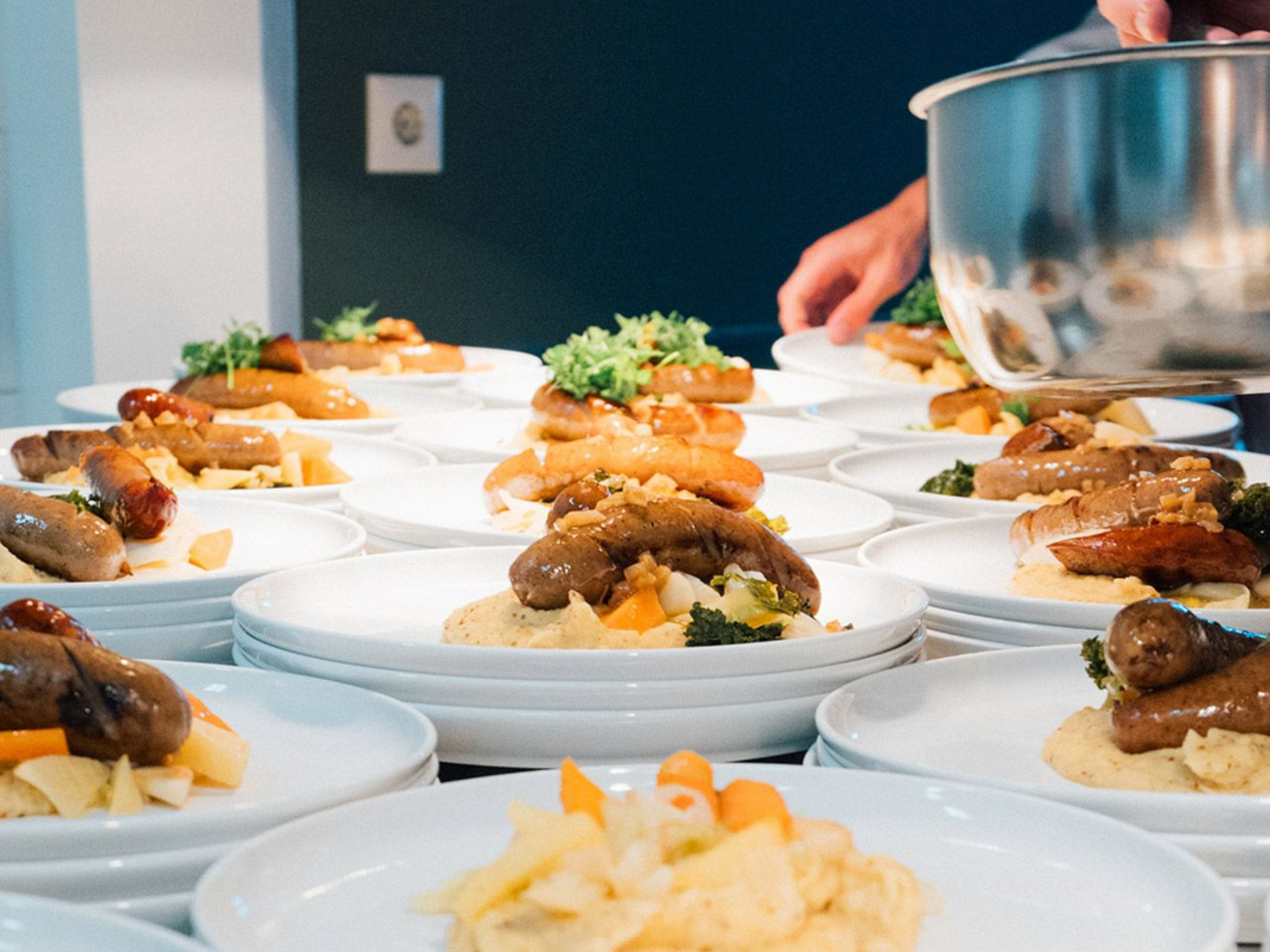 Tallerkener med pølse og potetmos blir servert på et kjøkken; en person heller fra en gryte.