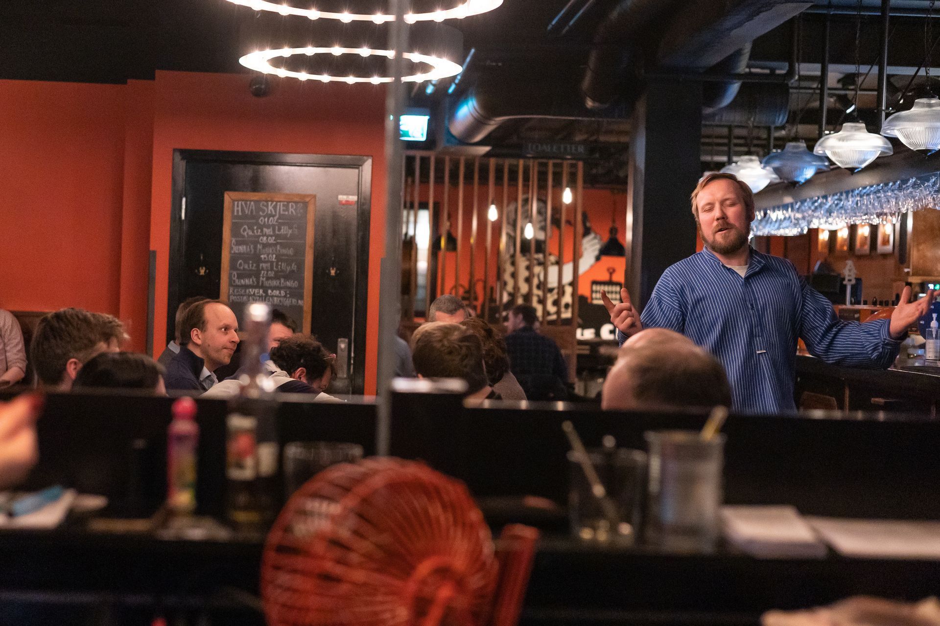 En svakt opplyst bar med en folkemengde. En mann på en scene med instrumenter. Arkadespill til høyre.