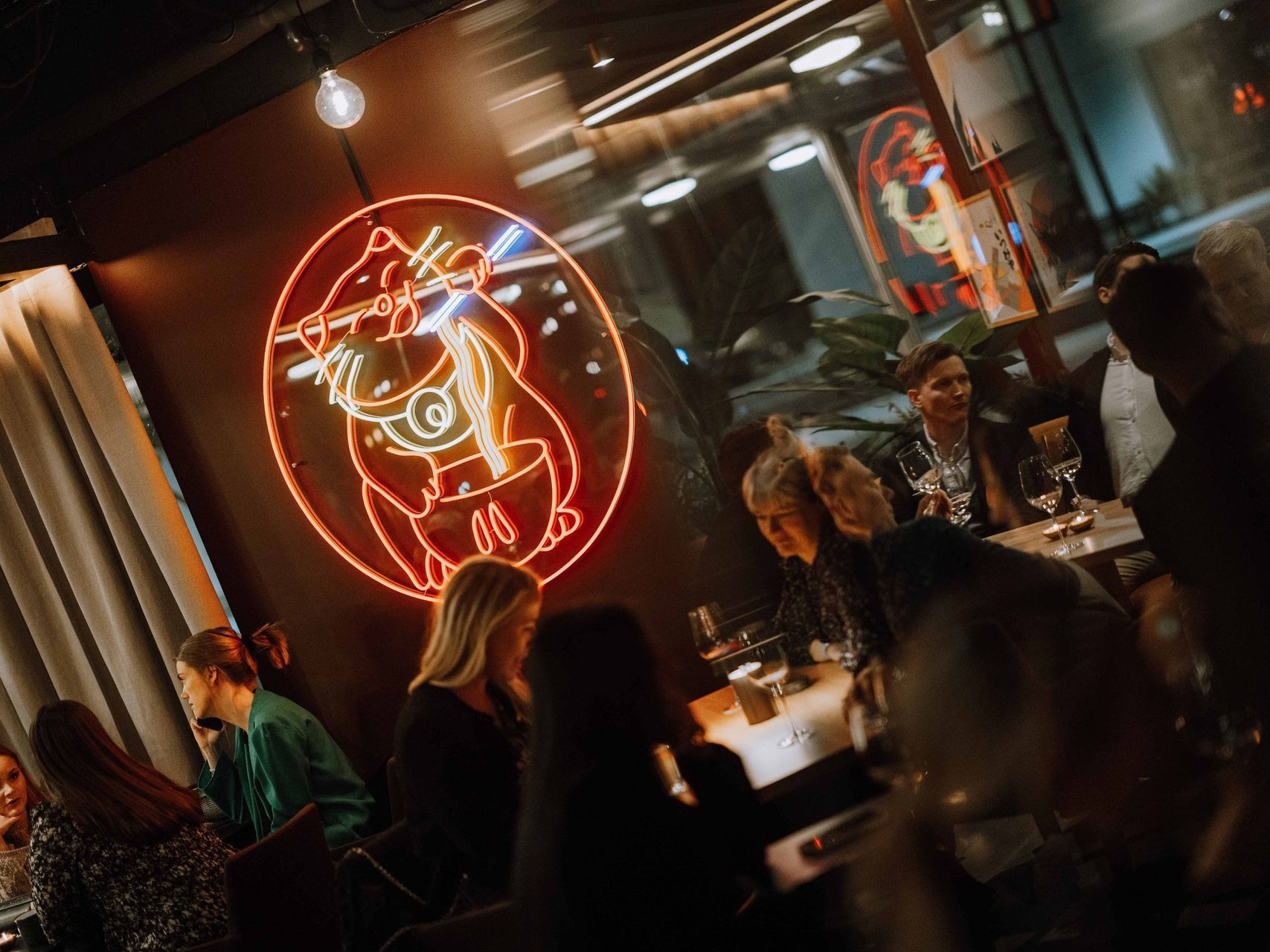 En gruppe mennesker sitter ved bord i en restaurant med et neonskilt på veggen.