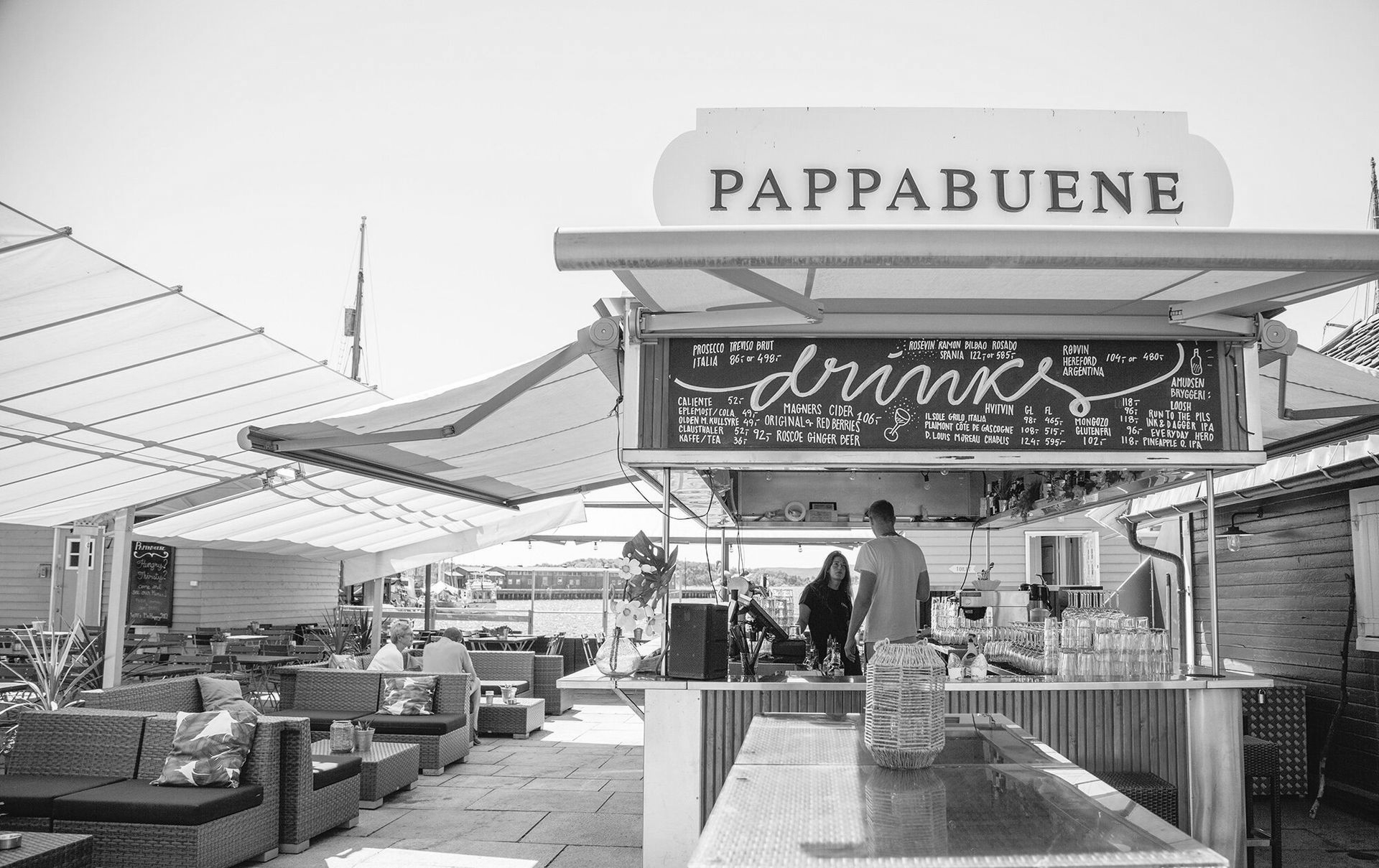 Utendørsbaren «Pappabuene», med drikkemeny og folk sittende ved bord under markiser.