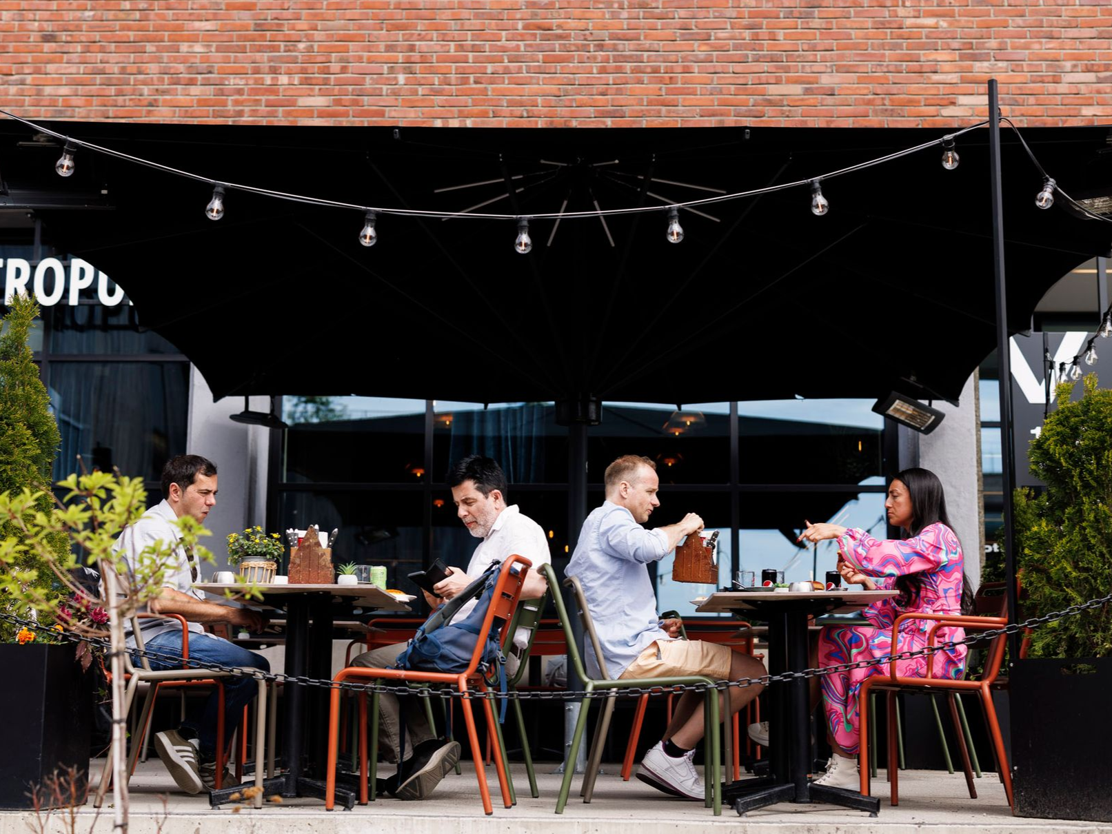 Folk sitter ved uteserveringsbord under en svart parasoll, spiser og snakker.