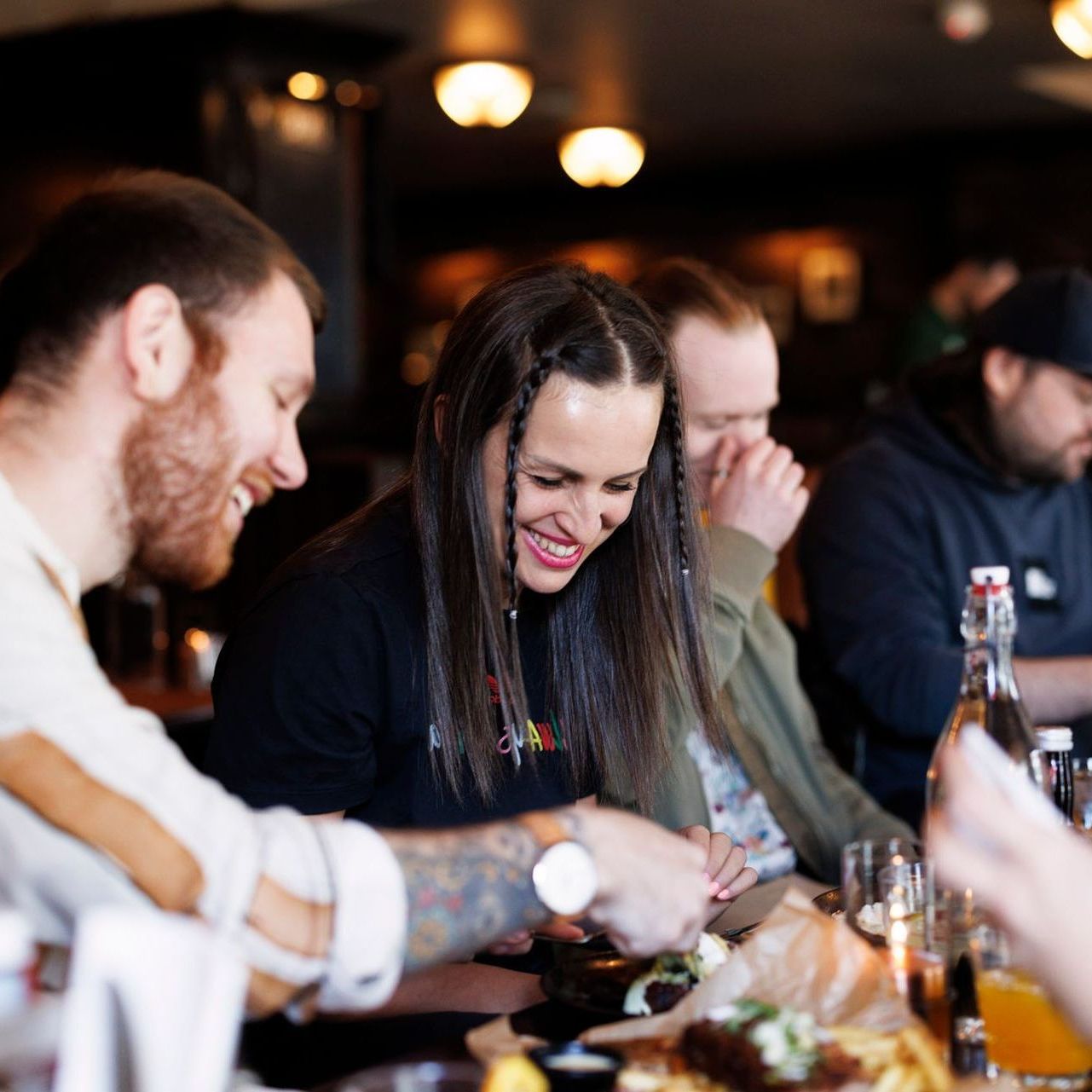 Folk ler rundt et bord på en restaurant. En kvinne smiler bredt. Mat og drikke er tilgjengelig.