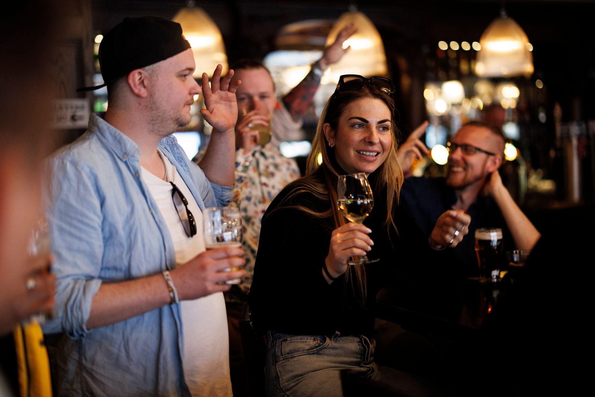 Gruppe mennesker i en bar, feirer og drikker. Kvinne med vinglass smiler.