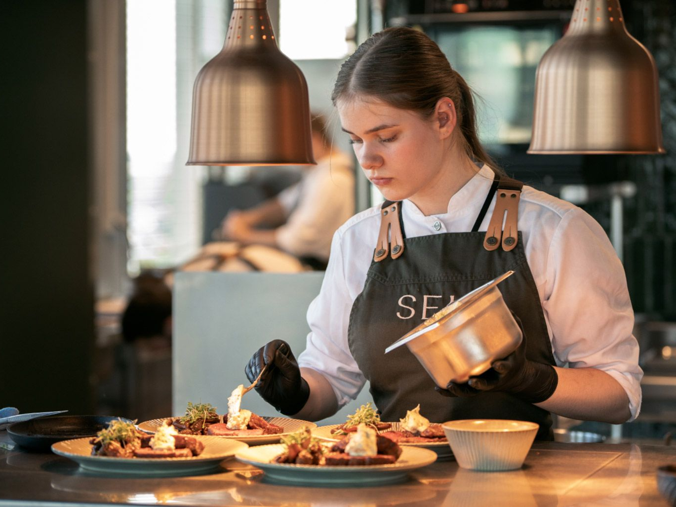 To bartendere i forklær i en bar: den ene sitter på disken med et glass, den andre ser på en klokke.