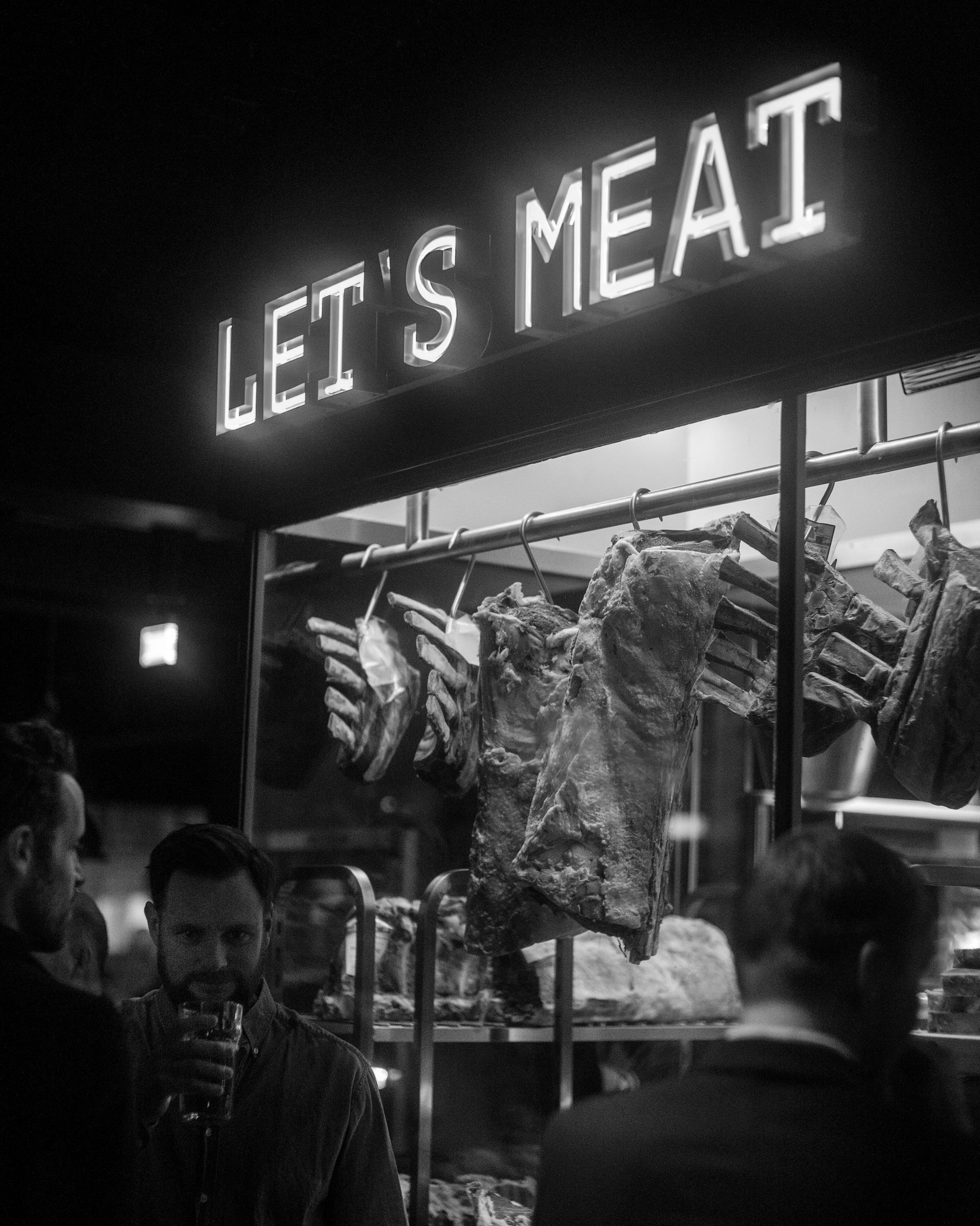 «Let's Meat»-neonskilt over et kjøttutstillingsskap i en mørk restaurant; folk i forgrunnen.