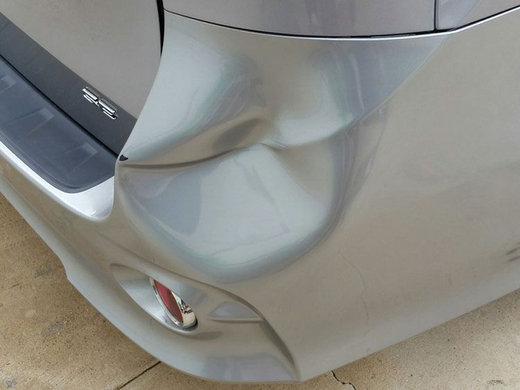 A silver car with a damaged bumper is parked on the side of the road