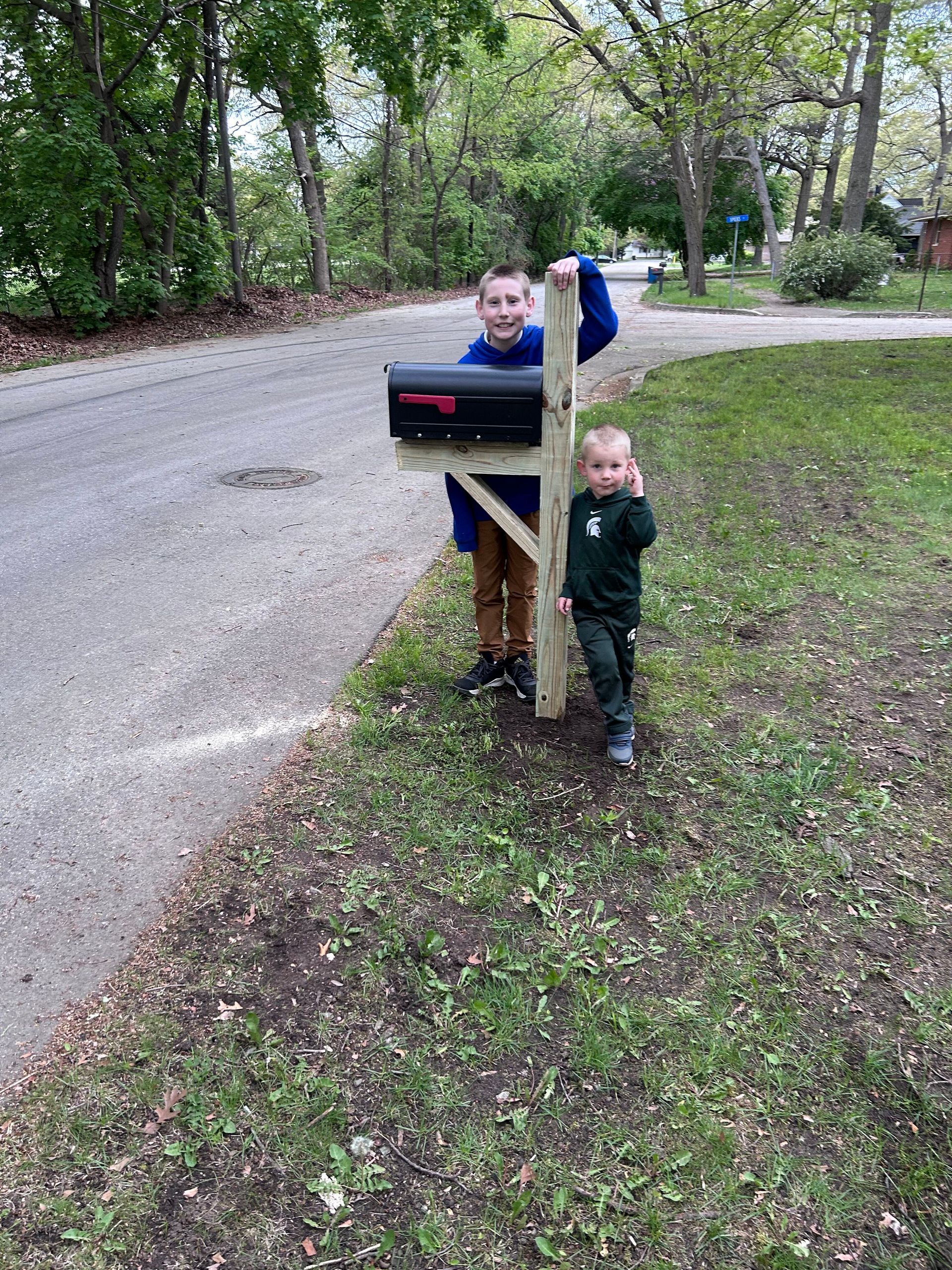 Two young boys are standing next to a mailbox on the side of the road.