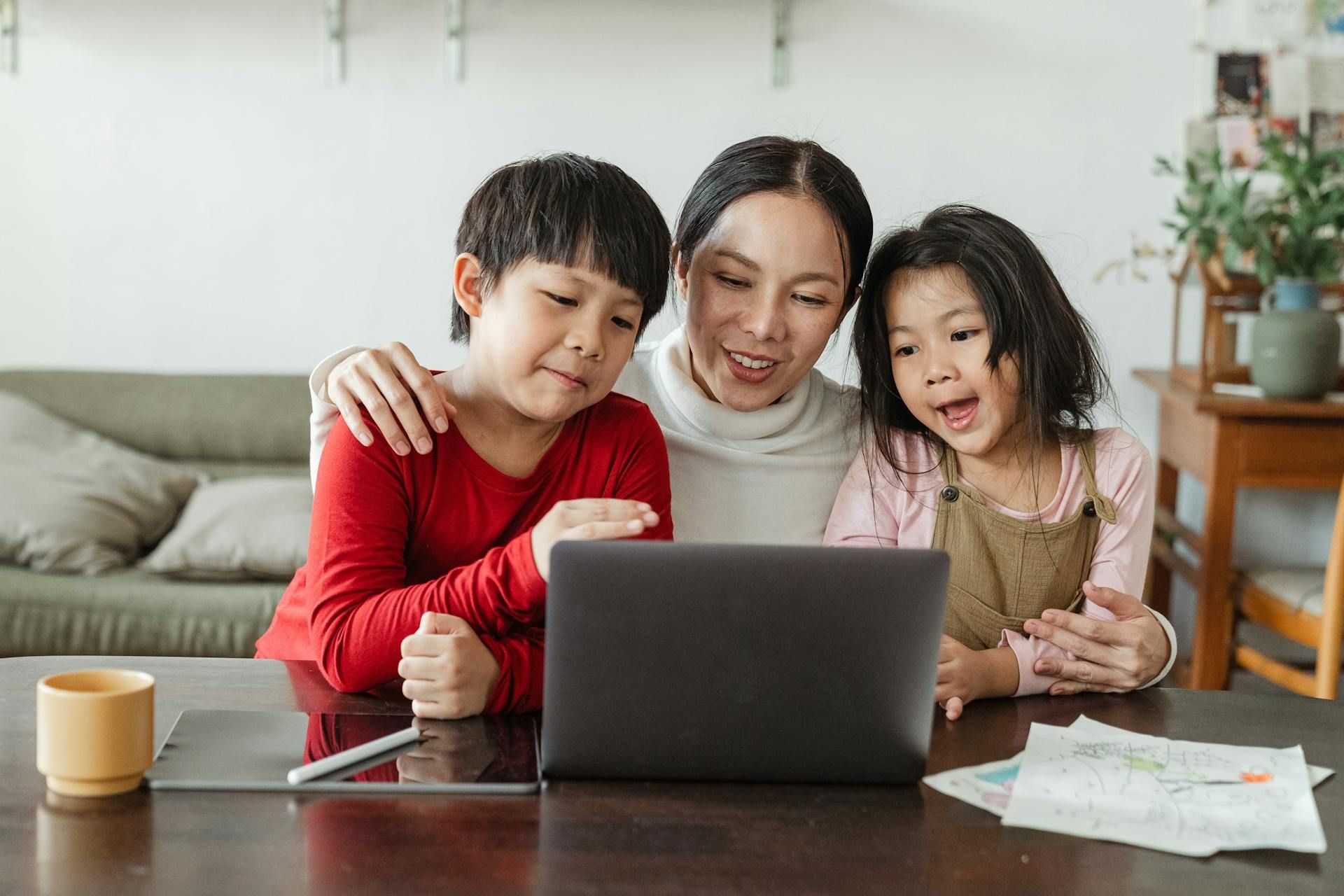 A smiling parent and two children sit at a table together, looking at a laptop screen in a bright, modern living room.