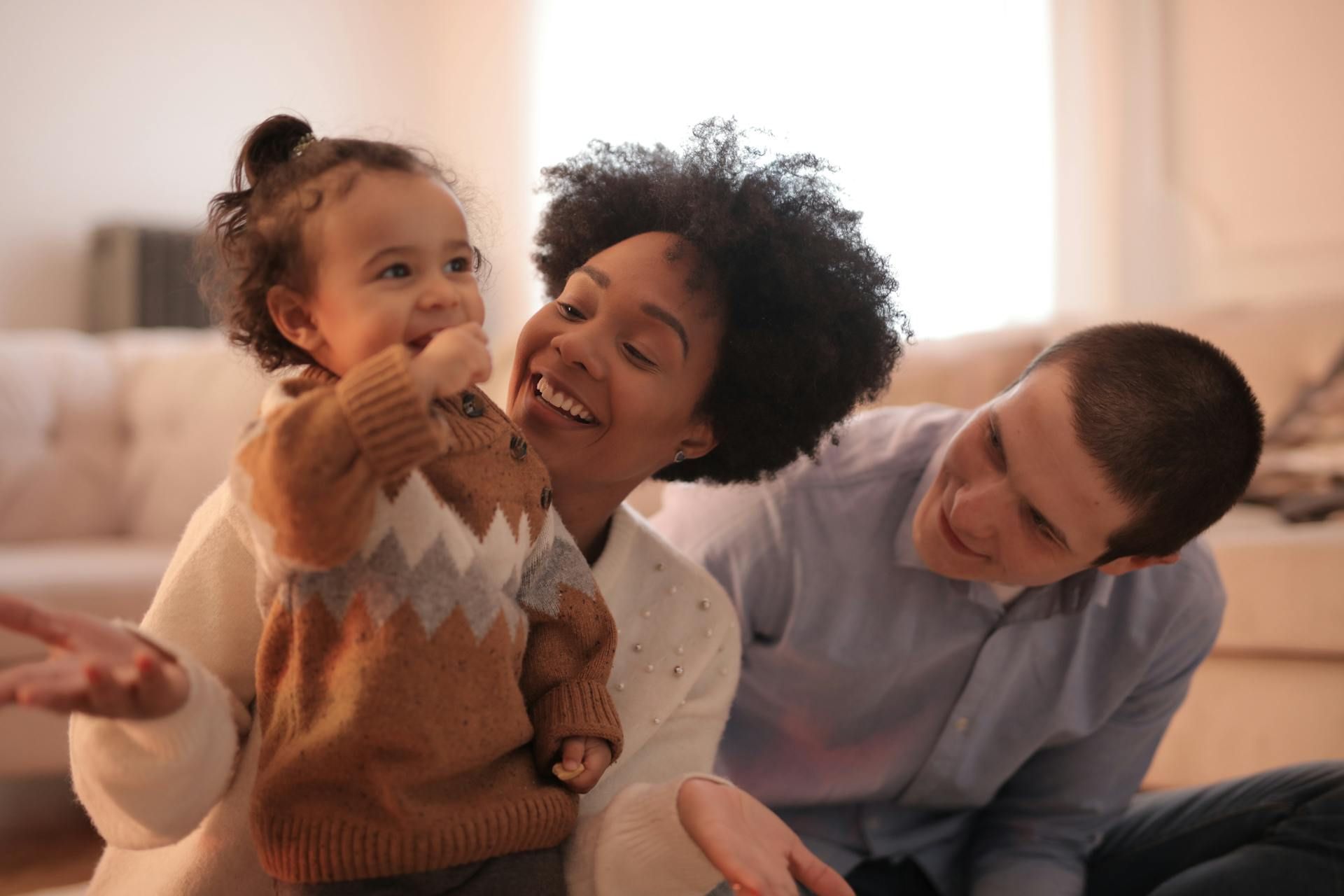 A toddler sits between two smiling adults in a sunlit living room, holding a small object to their mouth.