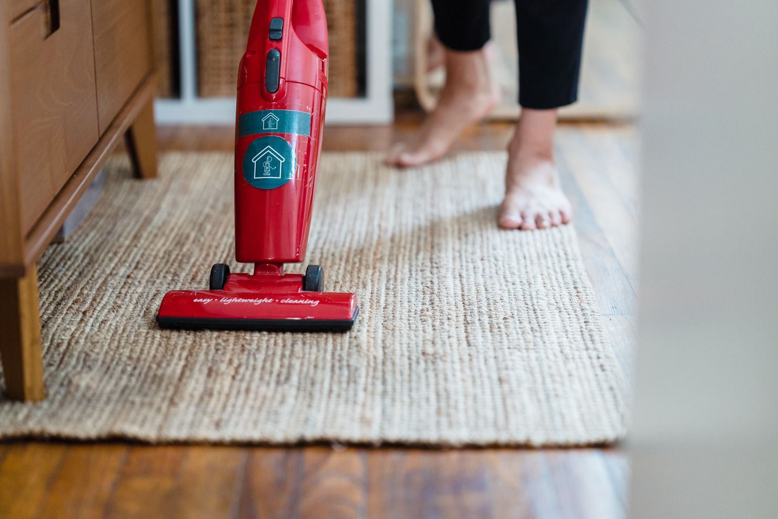 Red vacuum cleaner cleaning a beige rug, near a person's bare feet on a wooden floor.