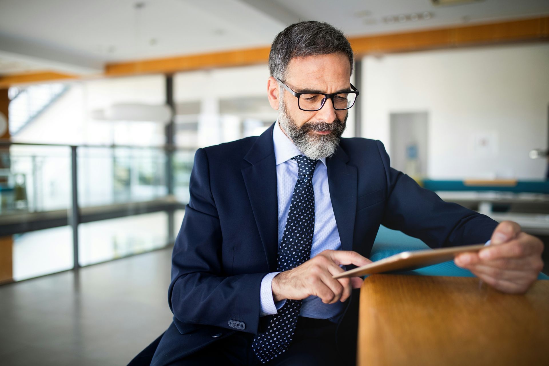 A professional in a suit and glasses sits at a table, focused while using a tablet in a bright office space.