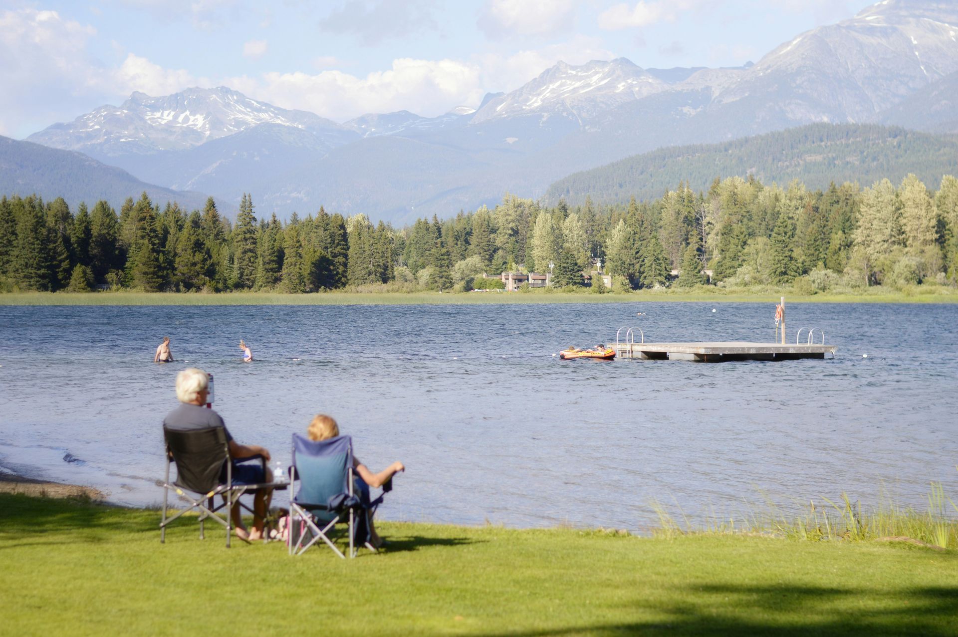Two people sit in folding chairs on a grassy lakeshore, looking out at a floating dock and distant, snowy mountains.