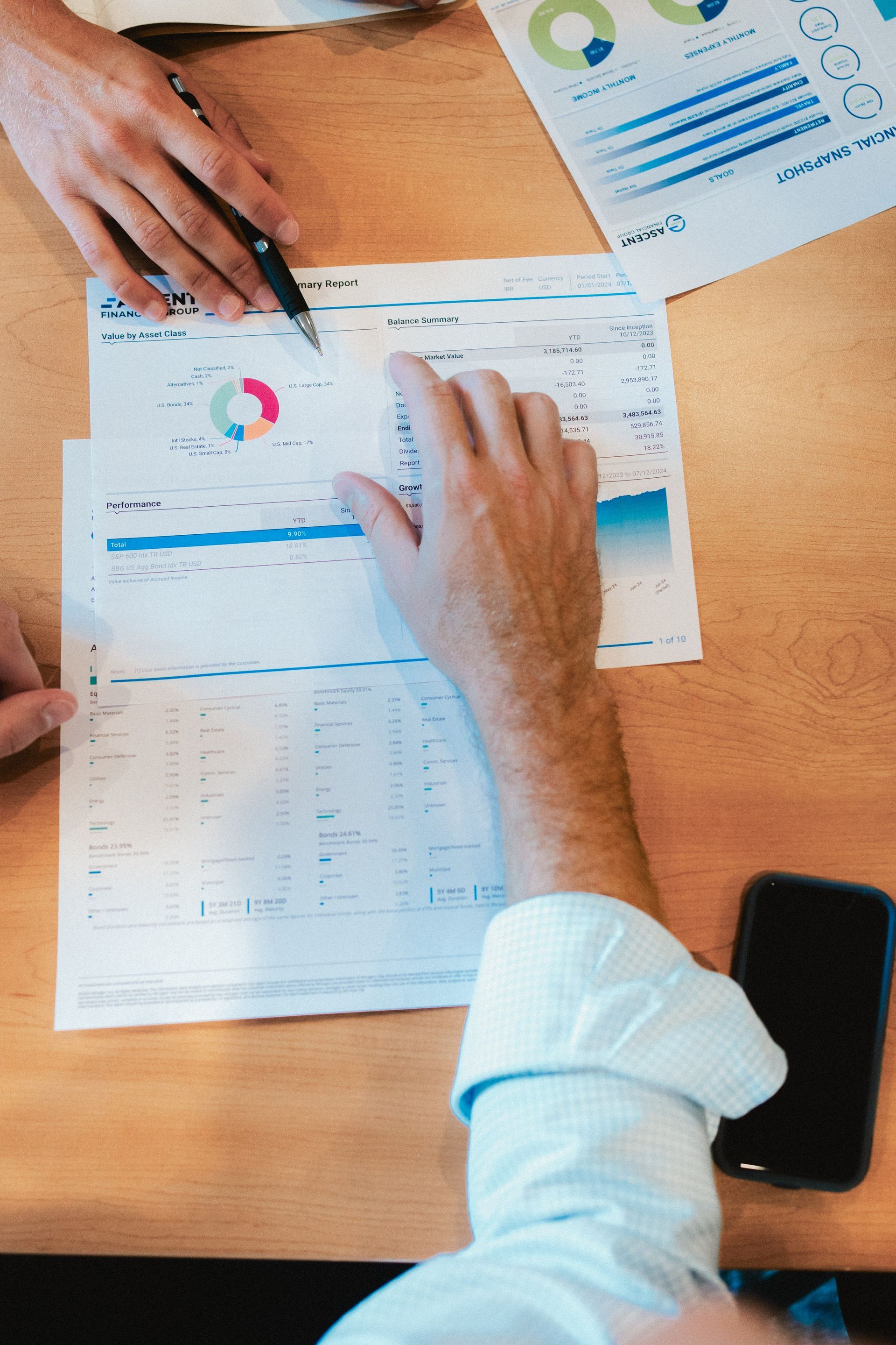 Hands pointing at printed business charts and data tables on a wooden table next to a smartphone.
