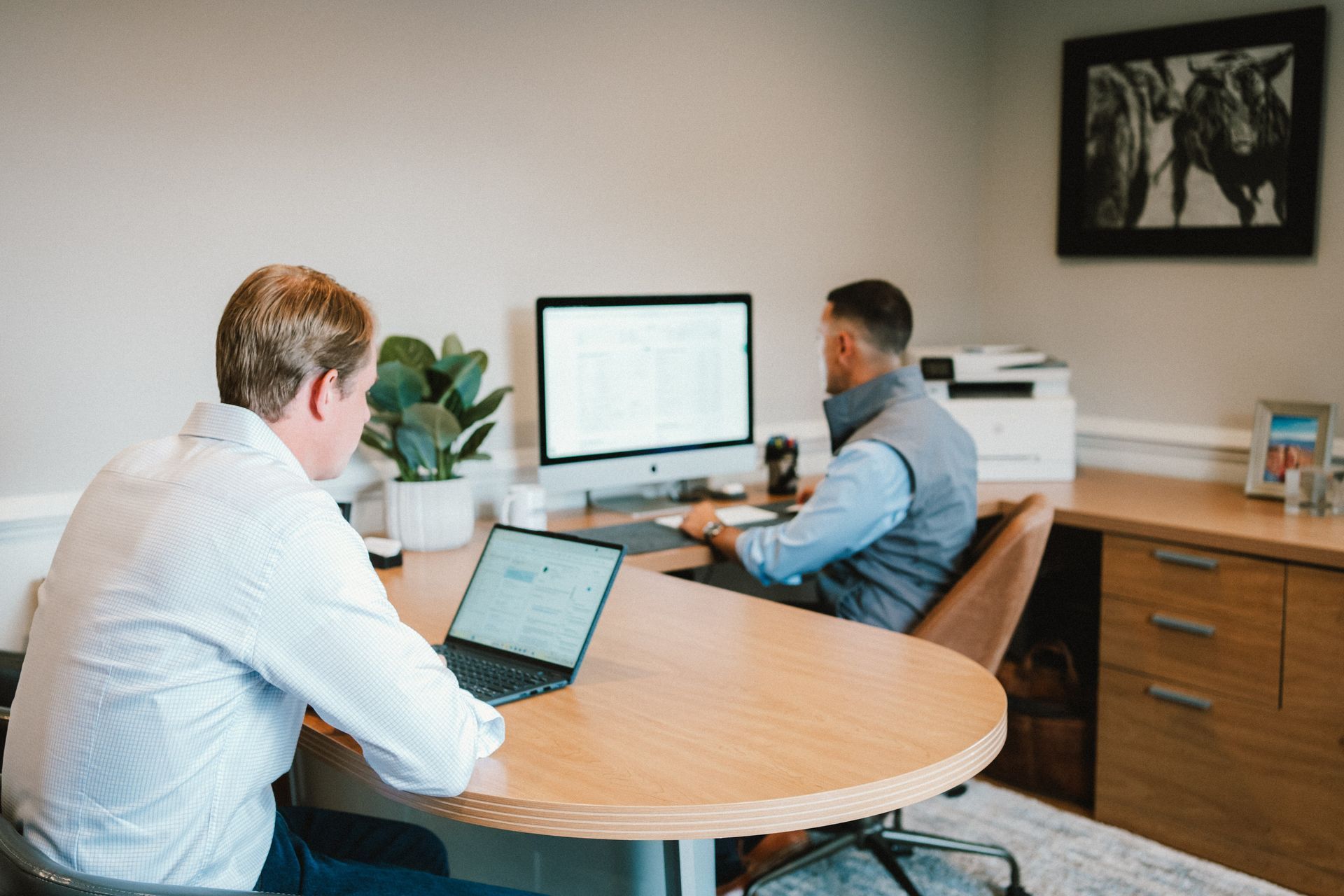 Two people sit at a desk in a professional office, one working on a laptop and the other on a desktop computer.