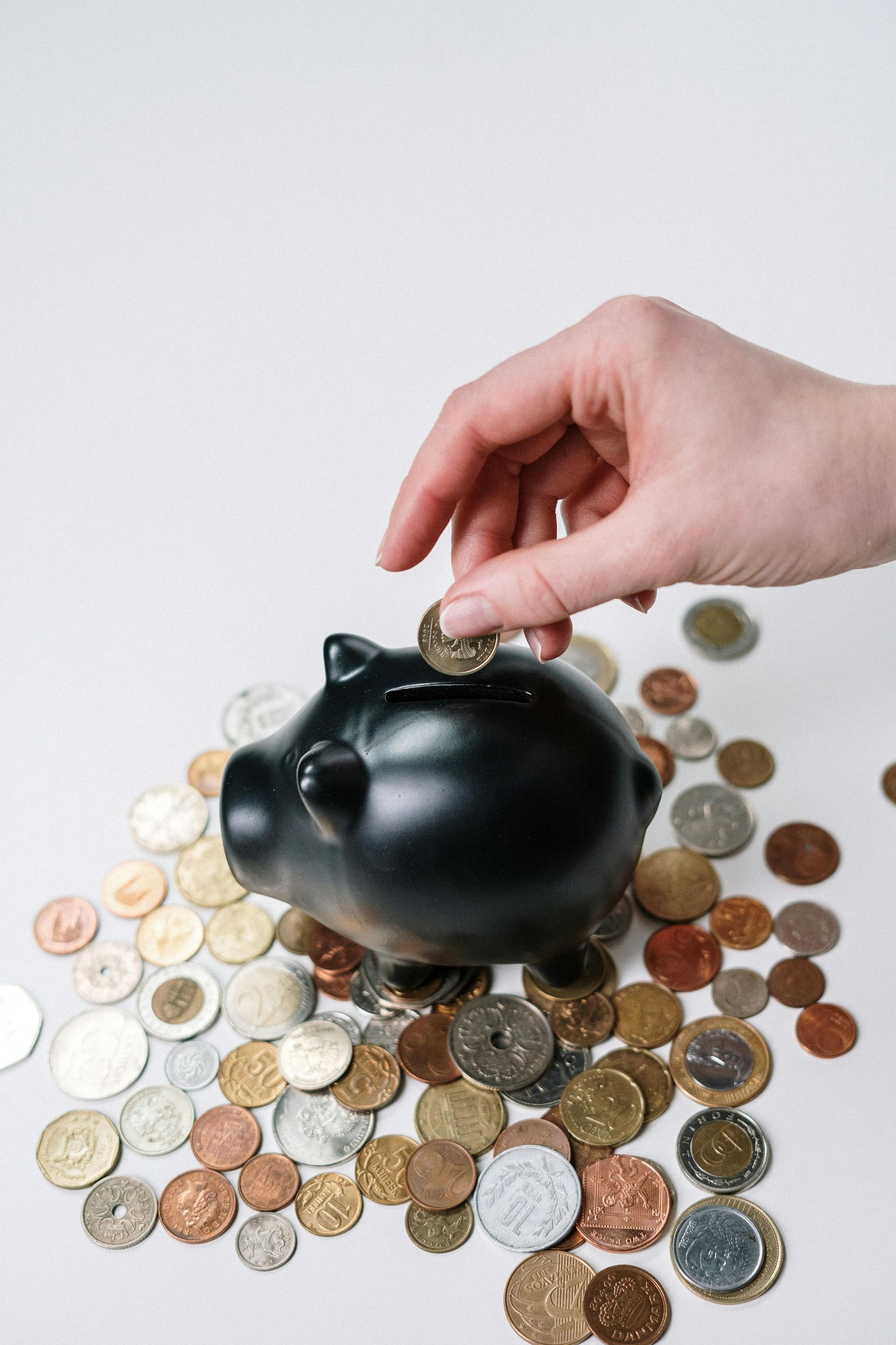 A hand places a coin into a black piggy bank surrounded by a pile of various coins on a plain white surface.