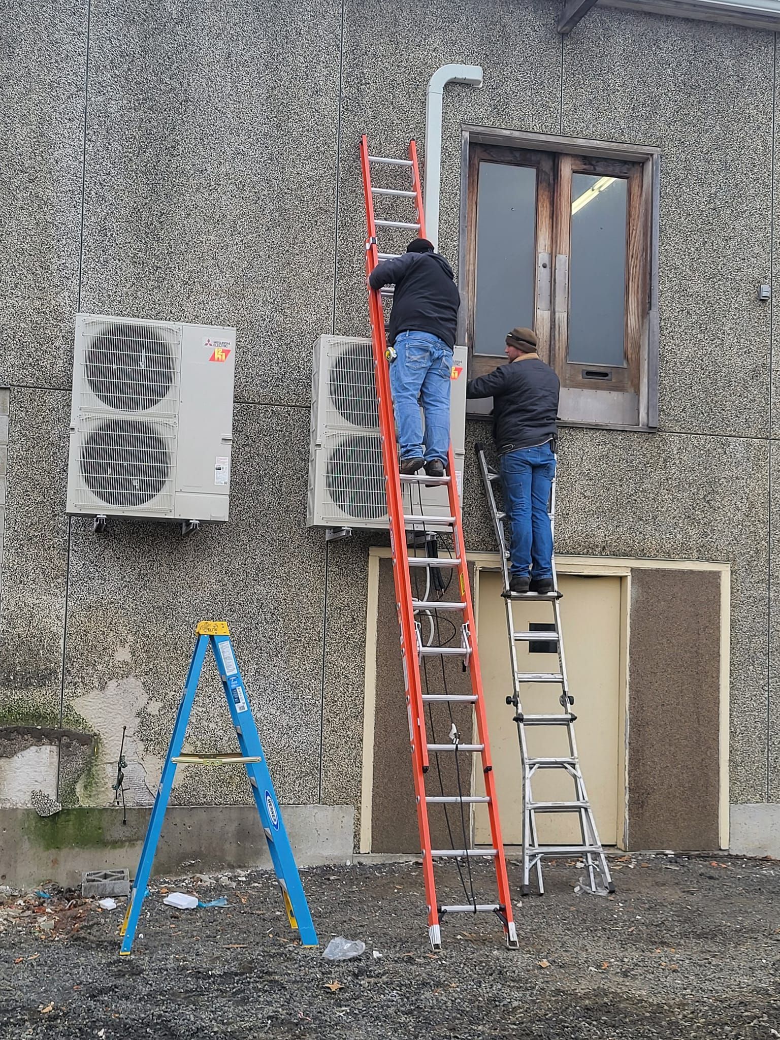 Two men are working on an air conditioner on the side of a building.