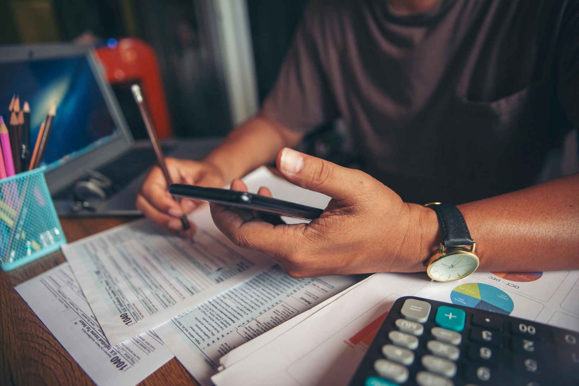 Person working at desk, holding phone and pencil, looking at tax forms. Calculator, laptop, and pens visible.