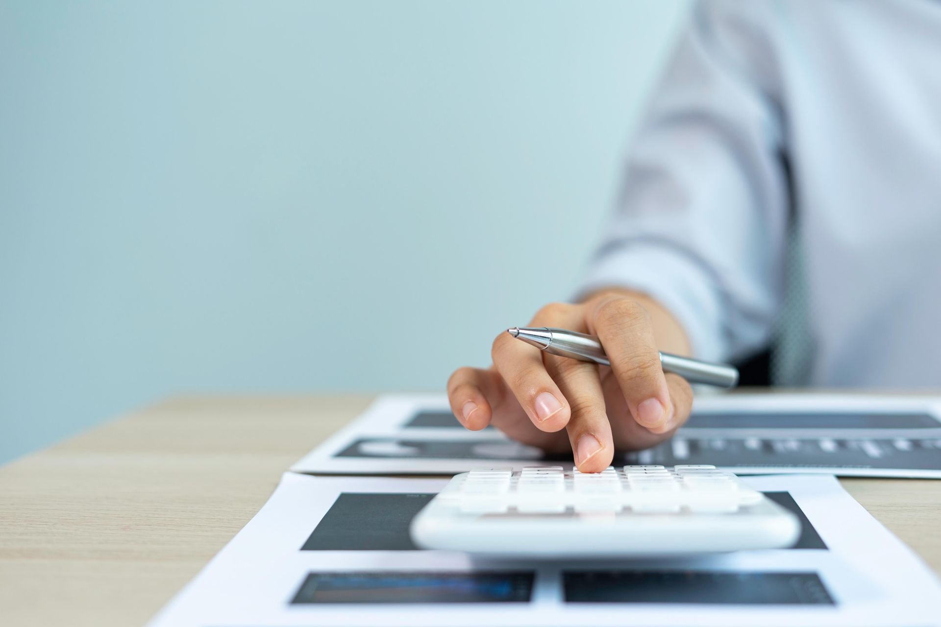 Person's hand using a calculator with a pen. Documents are on the desk, likely in an office setting.