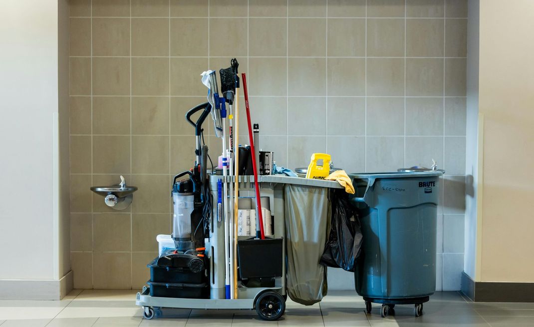 Cleaning supplies cart in a hallway, with a vacuum, mops, cleaning solutions, and a trash can, near a water fountain.