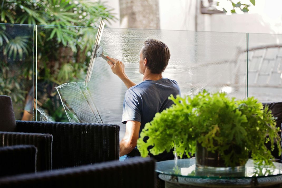 Man squeegeeing a glass panel. He's outdoors, near green plants and wicker chairs.