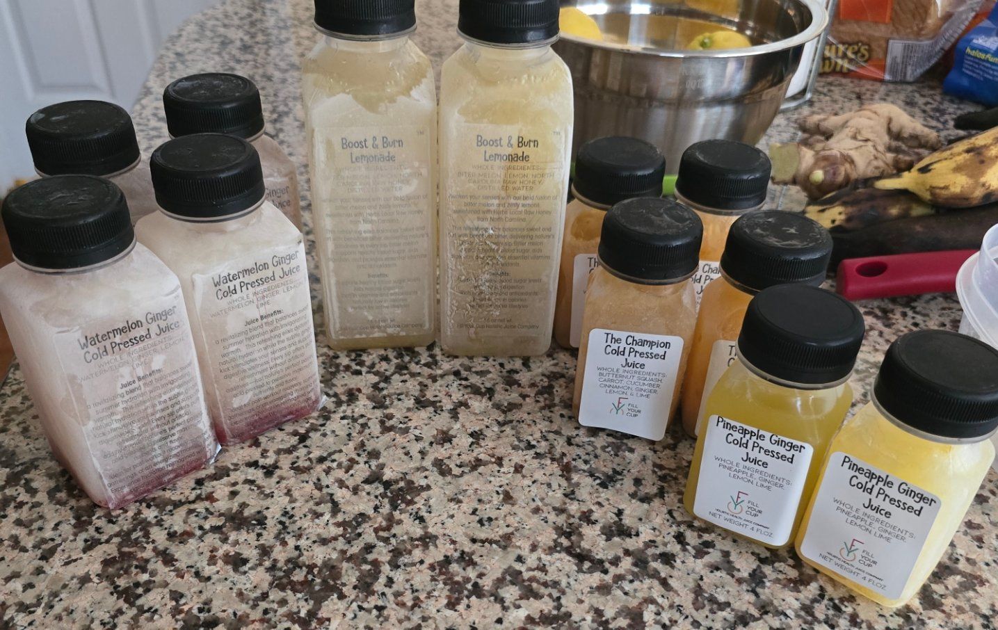 Bottles of fresh juice, various colors, on a countertop, with ingredients and a bowl in the background.