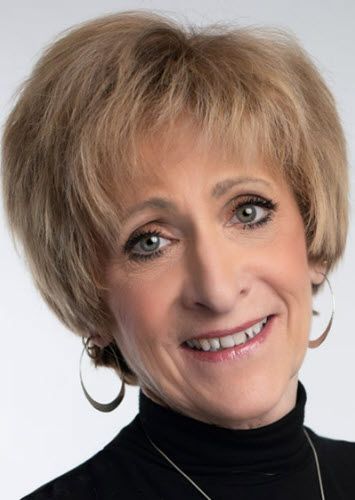 A woman, Grace Eisman, wearing a black turtleneck and earrings is smiling for the camera.