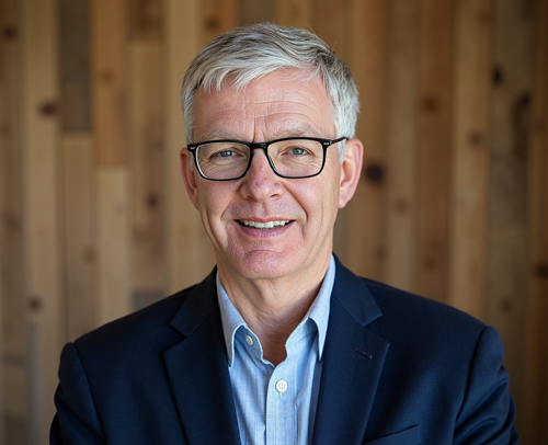 A man, Paul Evans, in a suit and glasses is smiling in front of a wooden wall.