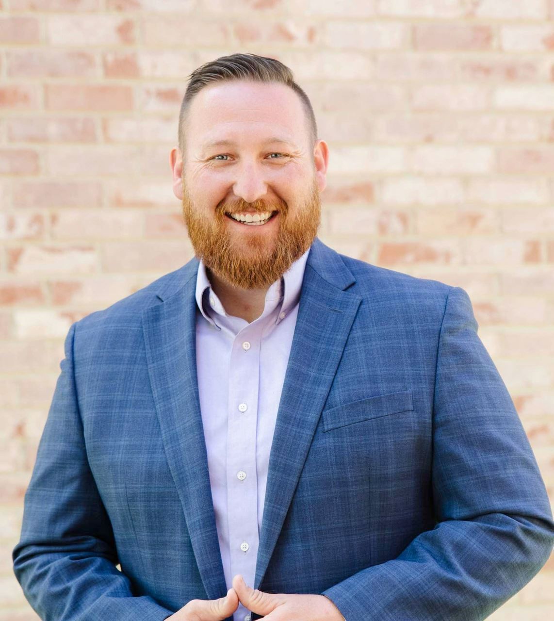 A man. Josh McReavy, with a beard is wearing a blue suit and smiling in front of a brick wall.