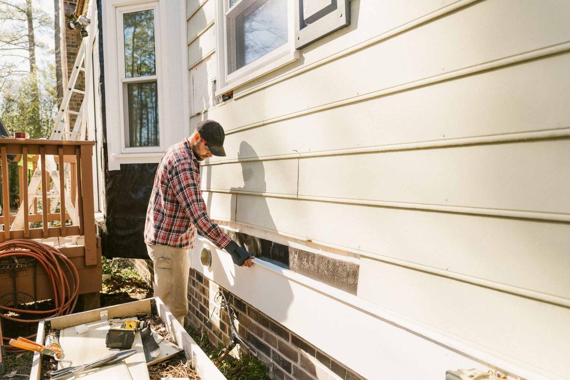 Man in a cap and plaid shirt repairs siding on a house exterior.